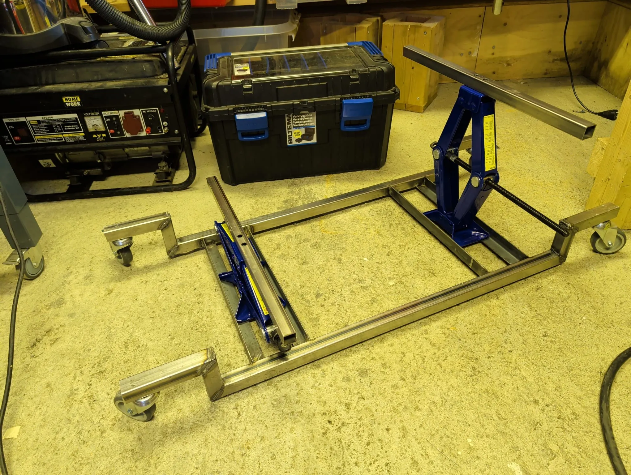 The drop dolly on the workshop floor before the drop: a welded ladder frame of 30 mm box section, a scissor jack on an angle shelf at each end, castors on mitred arms at the corners